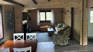 A cozy living room with a wooden ceiling, a sofa, a coffee table, and a dining area with a wooden table and white chairs. - Riverside cabin in the woods (Bracebridge)