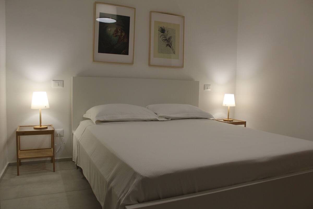 Ein ordentlich bezogenes Bett mit zwei Kissen, zwei Nachttischlampen und zwei gerahmten Bildern an der Wand.