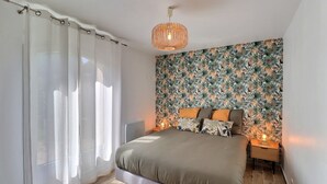 A bedroom with a bed, a nightstand, a lamp, a wall with a tropical leaf pattern, and sheer curtains. - La Maisonette Provençale (Ventabren)