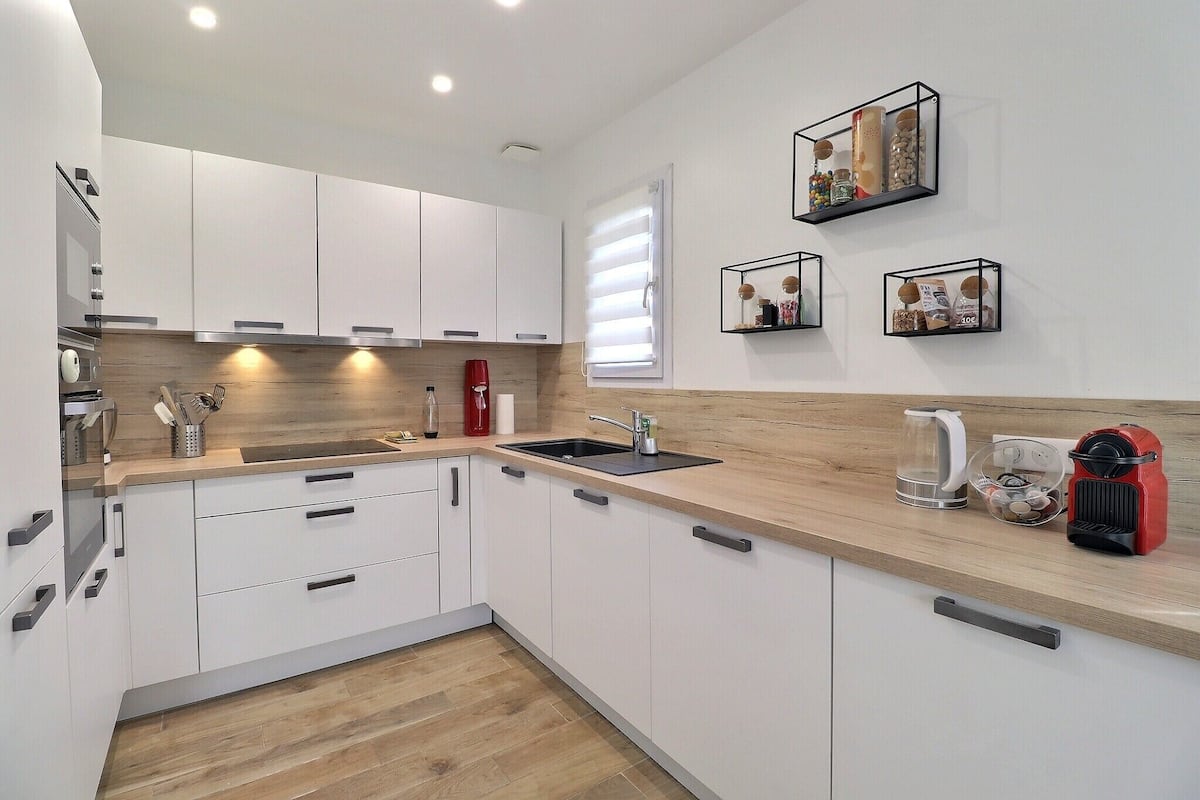 A modern kitchen with white cabinets, a wooden countertop, and a built-in oven. There are three black-framed wall shelves with decorative items.