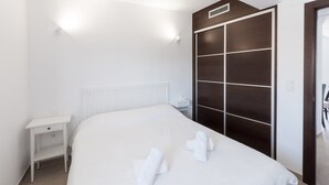 Cama bem arrumada com lençóis brancos e duas toalhas, mesa de cabeceira branca e guarda-roupa de madeira escura com porta espelhada.