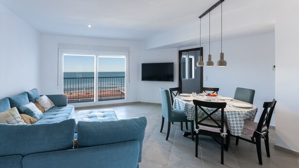 Uma sala de estar moderna com um sofá azul, uma mesa de jantar com cadeiras e uma TV fixada na parede.