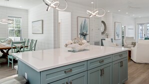 A modern kitchen with a large island, pendant lighting, and a dining area with a table and chairs. - NEW - Symphony! Sleeps 18! Pool! Golf Cart (Santa Rosa Beach)