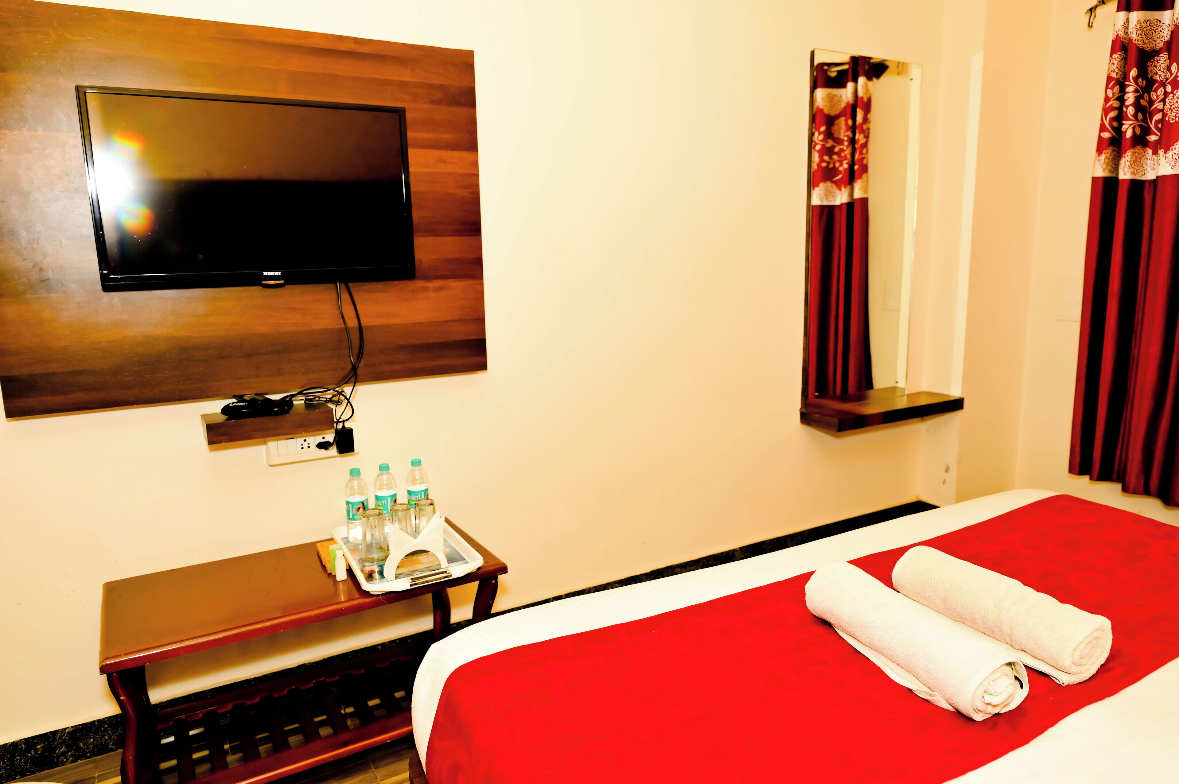 A hotel room with a flat-screen TV mounted on the wall, a bedside table with bottled water, a tray with a towel and a book, and a red bedspread with rolled towels.