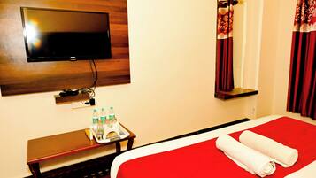 A hotel room with a flat-screen TV mounted on the wall, a bedside table with bottled water, a tray with a towel and a book, and a red bedspread with rolled towels.