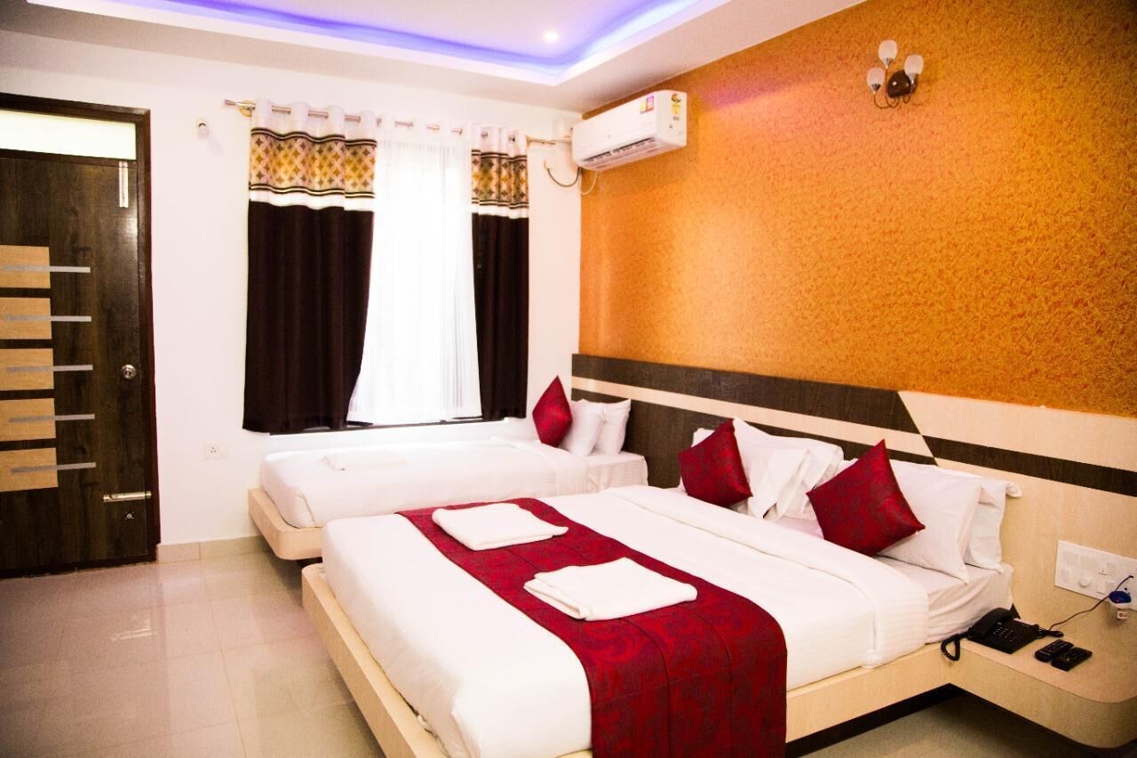 A hotel room with two beds, a wooden wardrobe, a window with curtains, an air conditioning unit, and a bedside table with a phone and remote controls.