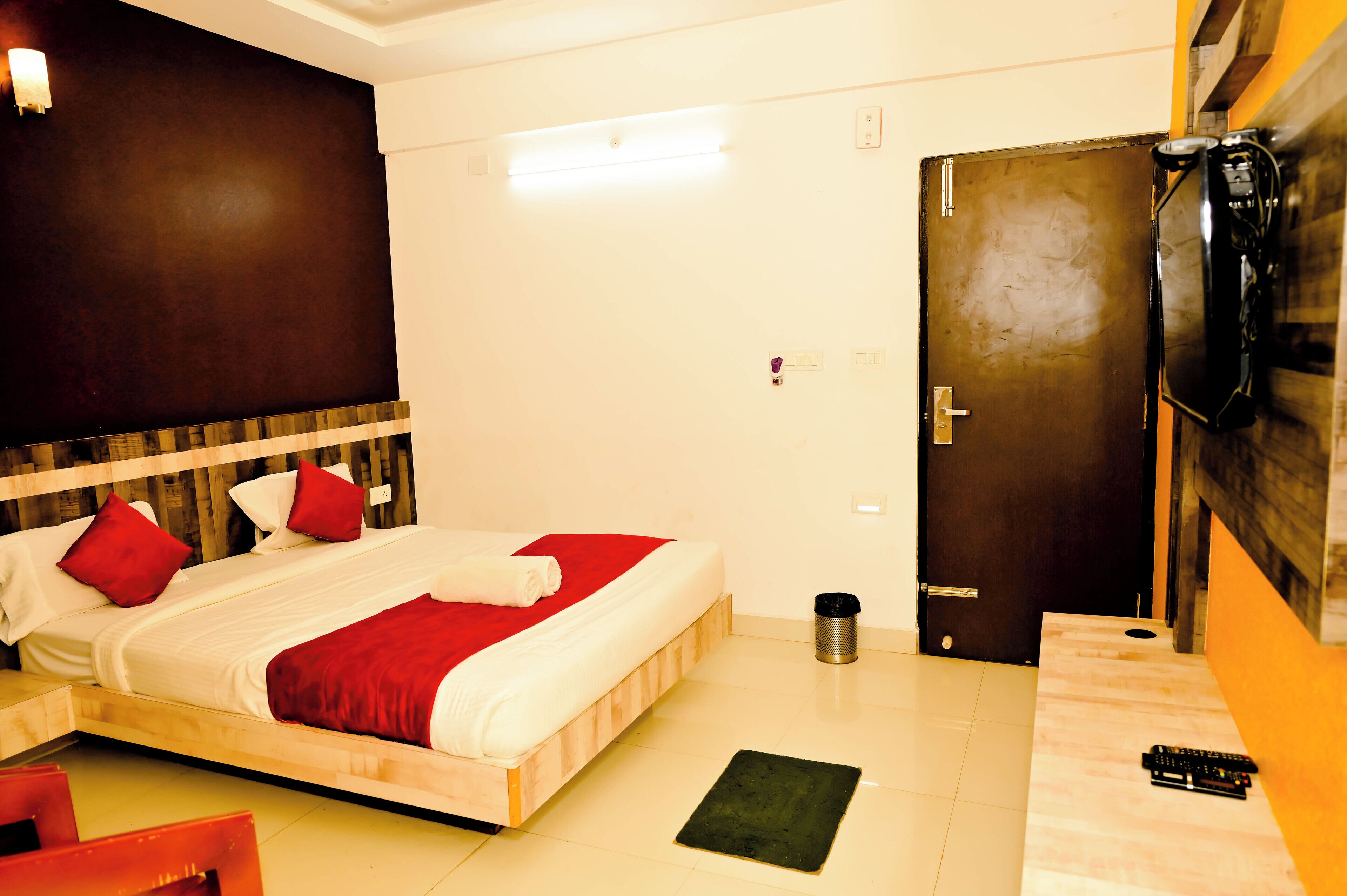 Habitación de hotel con cama, cabecera de madera, colcha roja y blanca, cesto de basura negro, mesita de noche de madera con control remoto y puerta con picaporte negro.