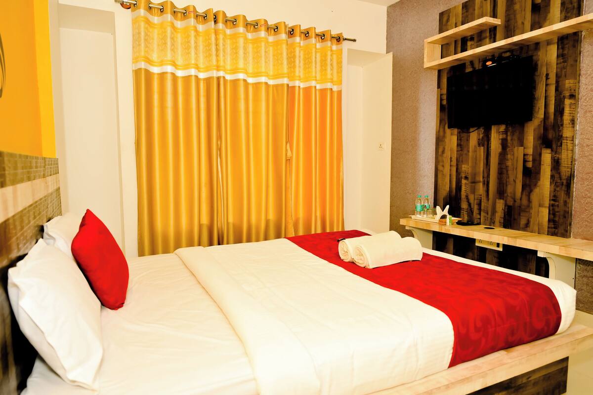 A hotel room with a bed, a red and white bedspread, a yellow curtain, a wooden wall with a TV, and a small shelf with toiletries.