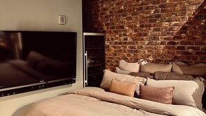 Een slaapkamer met een bakstenen muur, een flatscreen televisie, een bed met veel kussens en een houten vloer.
