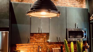 Een keuken met donkere kastjes, een bakstenen muur, een moderne hanglamp, een roestvrijstalen koelkast en een magnetron.