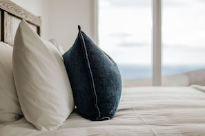 Ein Bett mit zwei Kissen, ein weißes und ein dunkelblaues, in einem Zimmer mit Blick durch ein Fenster.