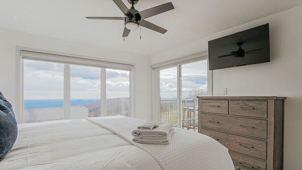 A bedroom with a large bed, a dresser, and a ceiling fan. There are two windows providing a view of the outdoors. - Mountain View Vista: Resort Condo w/ Views (Wintergreen Resort)