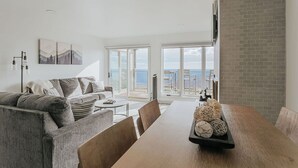 A modern living room with a large wooden dining table, grey sofa, and a view of the sea through large windows. - Mountain View Vista: Resort Condo w/ Views (Wintergreen Resort)