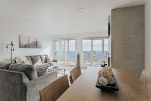 Un salon moderne avec une grande table à manger en bois, un canapé gris et une vue sur la mer grâce à de grandes fenêtres.