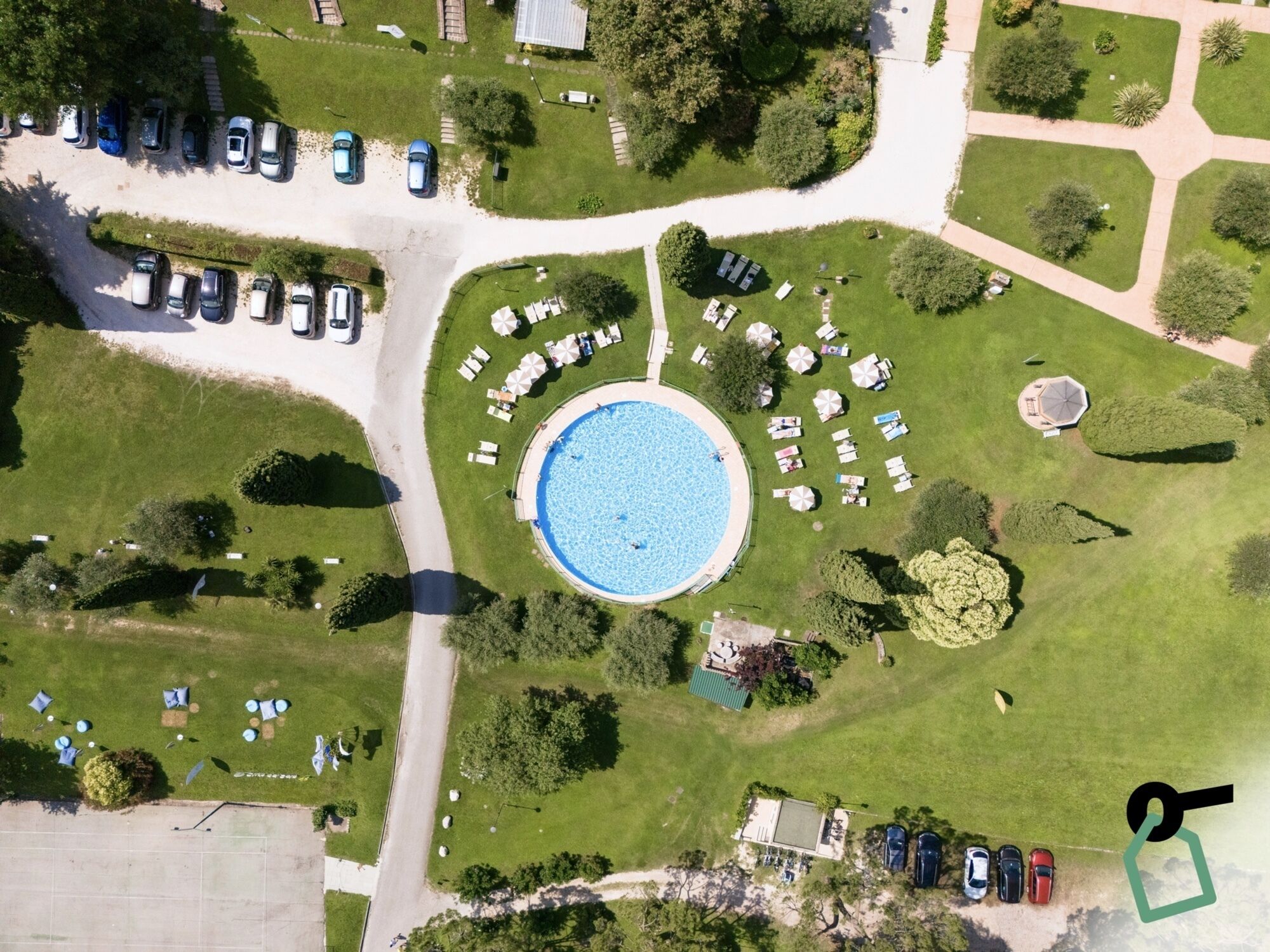 An aerial view of a resort with a central swimming pool, arranged outdoor seating, and a parking area.