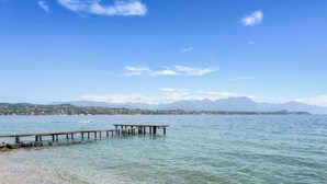 Un lago transparente con un muelle de madera que se adentra en el agua, rodeado de montañas y un pueblo distante.