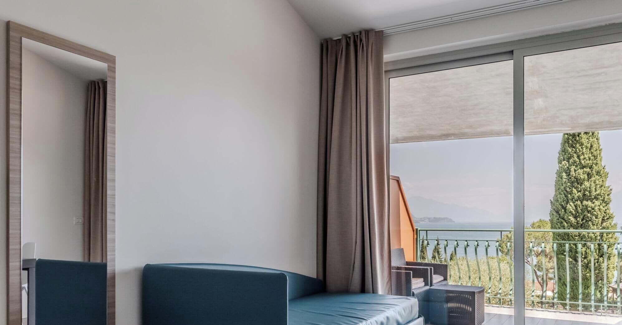 Quarto de hotel com sofá, poltrona, uma grande janela com vista para o mar e varanda com grade.
