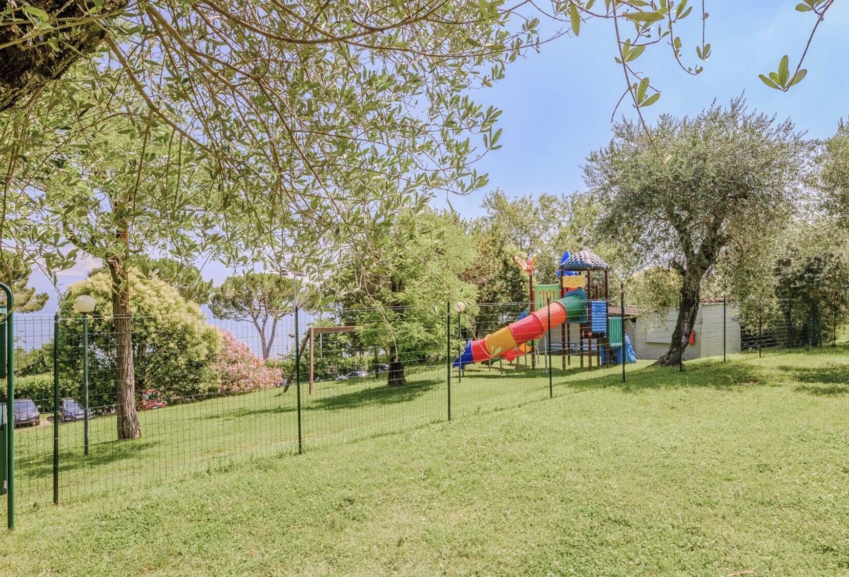 Une aire de jeux avec un toboggan et des structures d’escalade, entourée d’une clôture et d’arbres.