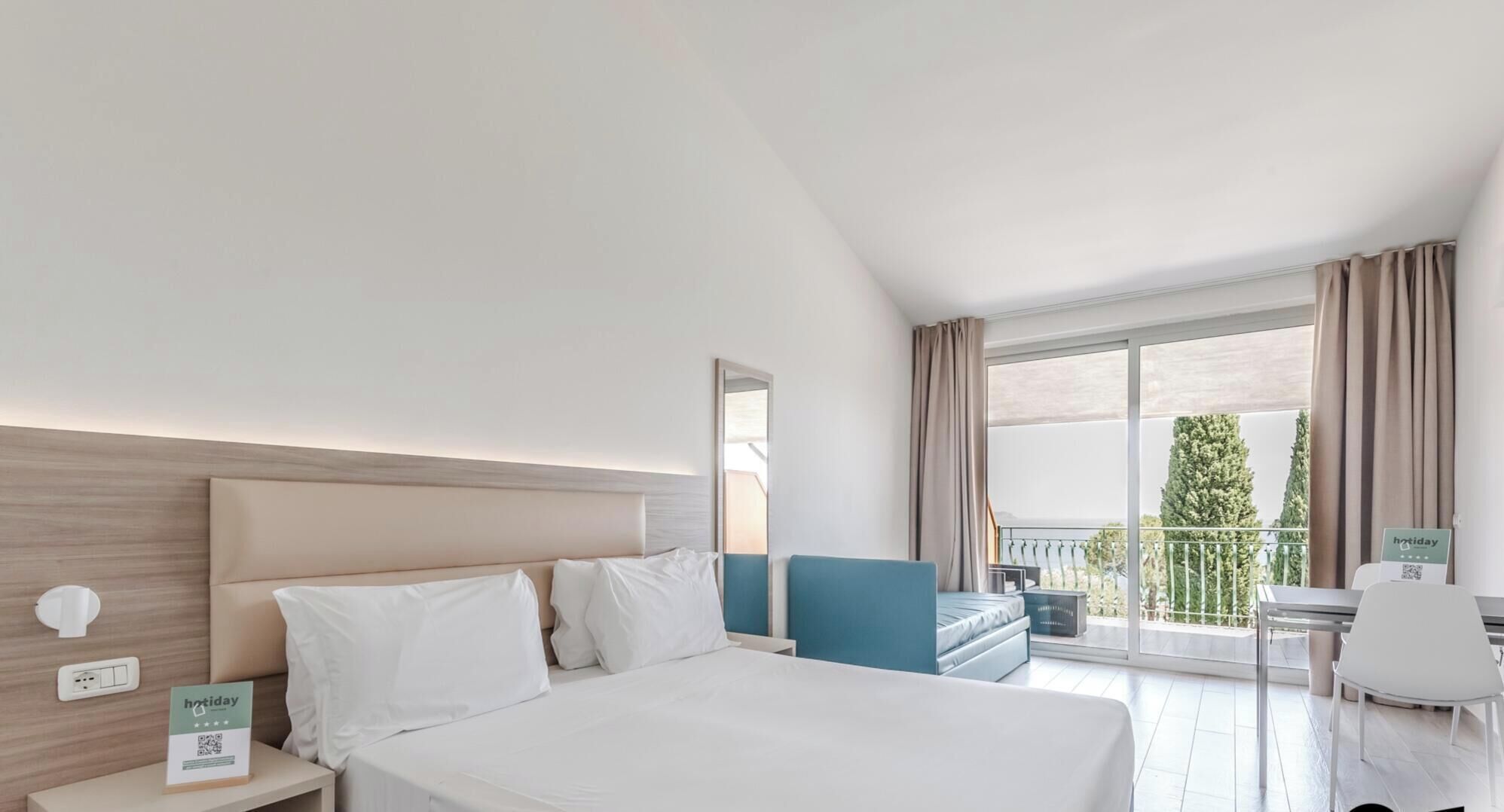 Habitación de hotel con una cama grande, un escritorio con silla, un balcón con vistas y un cartel con el nombre del hotel.