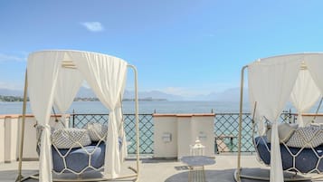 Zwei Betten mit weißem Moskitonetz und gemusterten Kissen, ein kleiner runder Tisch und Blick auf das Meer und die Berge.