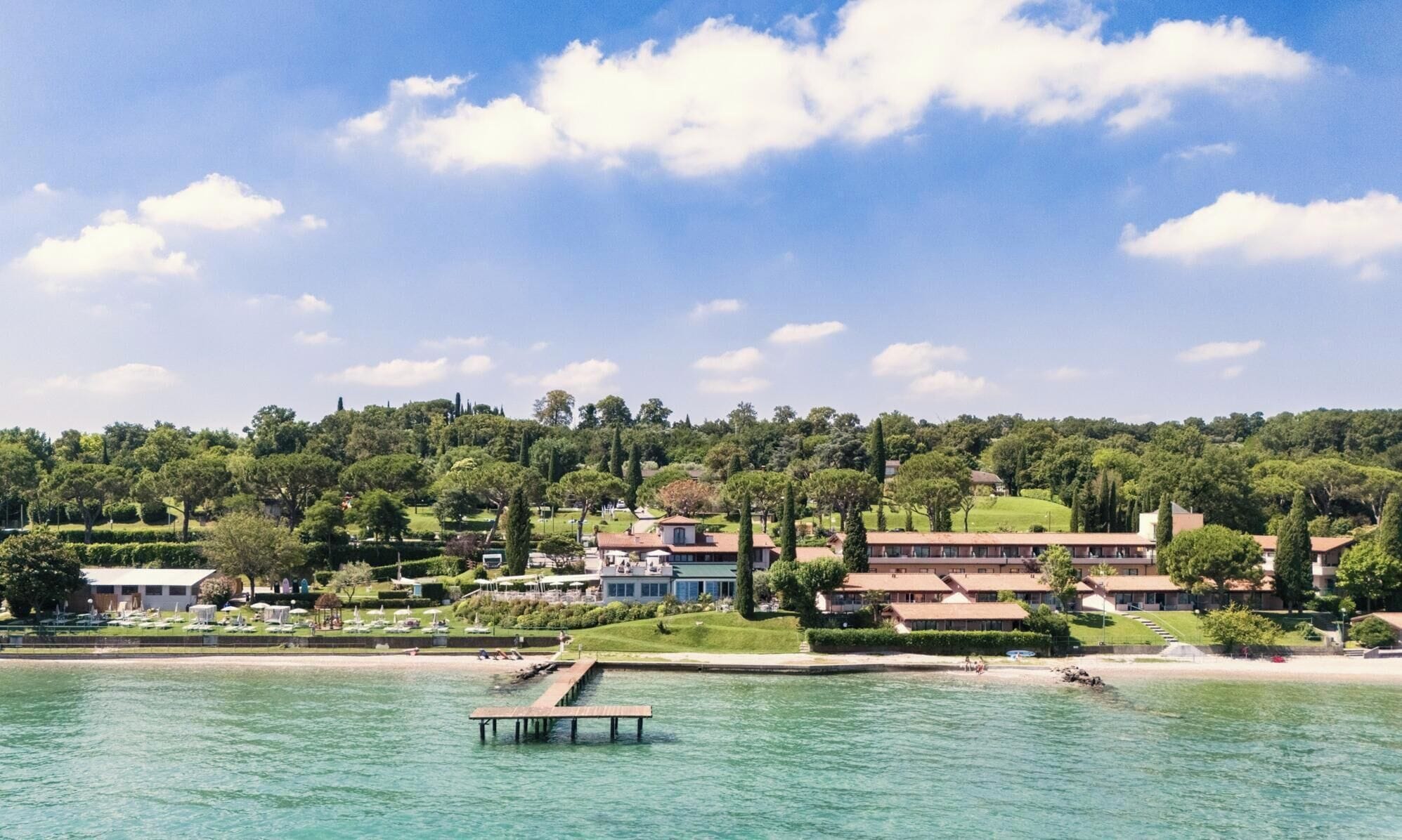 Un hotel frente al mar con un muelle que se adentra en el agua, rodeado de exuberante vegetación y colinas al fondo.