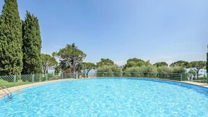 Una piscina azul cristalina rodeada de una cerca verde, con árboles y un cielo despejado al fondo.