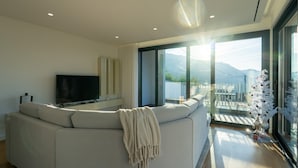 A modern living room with a large L-shaped sofa, a flat-screen TV, and a balcony with a view of the mountains.