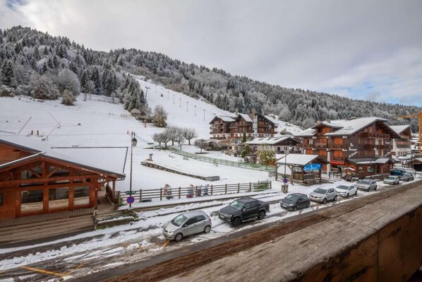 A snowy mountain village with wooden buildings, a ski slope, and parked cars. - Resort center, 2nd floor, balcony, tv, 30m², Morzine (Morzine)
