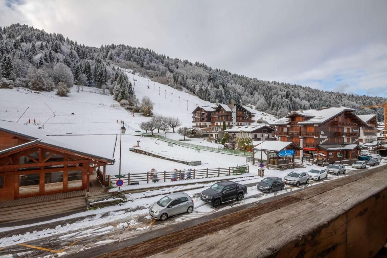 A snowy mountain village with wooden buildings, a ski slope, and parked cars.