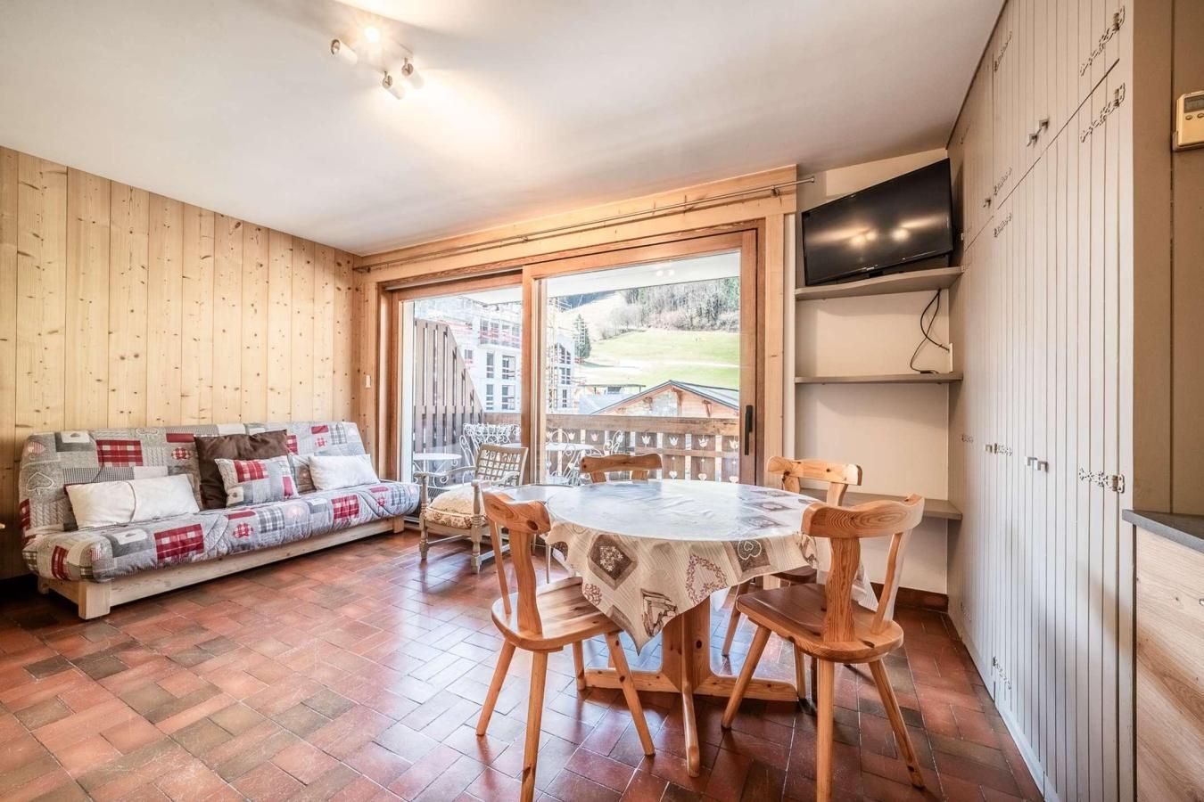 A cozy room with a wooden floor, a sofa with patterned cushions, a round table with chairs, and a large window offering a view of buildings and a fence.