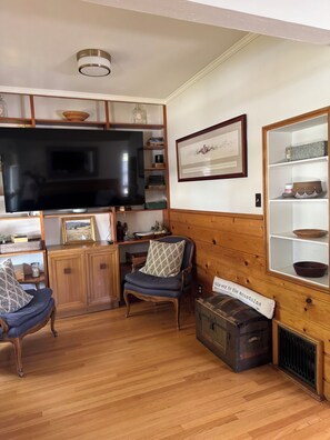 A living room with a television, wooden cabinets, a chair, a cushioned bench, and a vintage trunk.