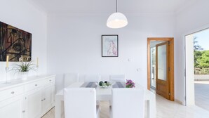 Un comedor moderno con una mesa blanca, sillas e iluminación colgante. Hay un cuadro en la pared y una puerta que da al exterior.
