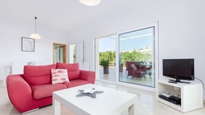 Una sala de estar moderna con un sofá rojo, una mesa de centro blanca, un televisor de pantalla plana sobre un mueble blanco y un amplio ventanal de vidrio que da a un patio exterior.