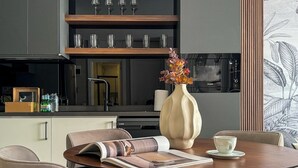 Un comedor moderno con una mesa de madera, sillas color beige, un florero con flores y una revista.