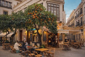 An outdoor cafe with tables and chairs, a tree with oranges, and a building with a bar named 'ATANCHON BAR'. - Modern apartment with balcony and WiFi in Sevilla Centro (Sevilla)