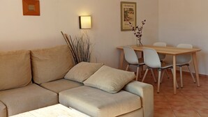A living room with a beige sofa, a dining table with chairs, and a wall-mounted light. - Burgit - Apartment In Es Cap Des Moro. Free Wifi (Porto Petro, Illes Balears)