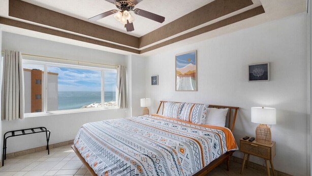 Une chambre à coucher avec un lit, un ventilateur de plafond, une fenêtre donnant sur la mer et une table de chevet avec une lampe.
