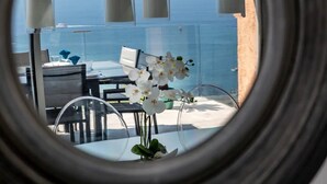 A view through a circular window of a dining area with white orchids, a table, chairs, and a view of the ocean.