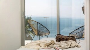 A bedroom with a large window, two wicker chairs, a bed with a blanket and a rolled-up magazine, and a handbag on the bed.