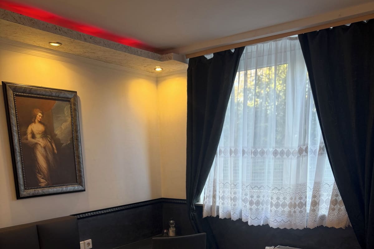 A hotel room with a bed, a desk, a chair, a radiator, and a window with curtains.