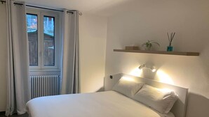A hotel room with a bed, a window with curtains, a radiator, a shelf with decorative items, and wall-mounted lights. - Spazio Perlasca apt1 - Cernobbio Como Lake (Cernobbio)