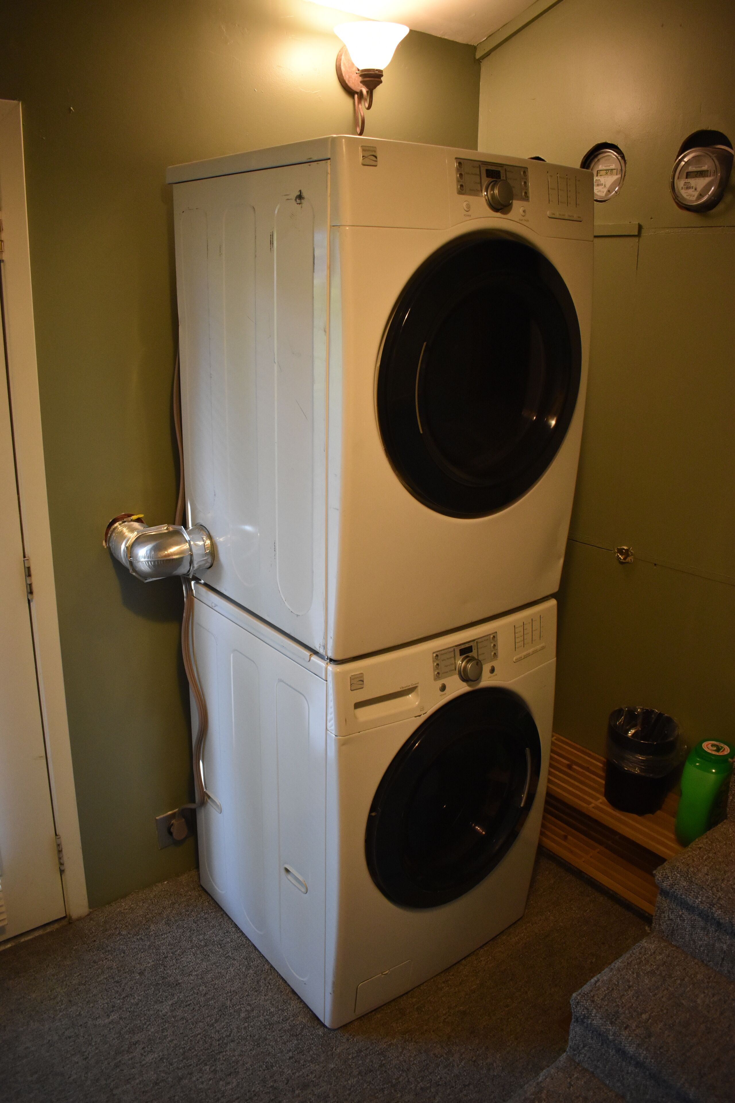 A stacked washer and dryer in a room with green walls, a wooden shelf, and a trash bin.
