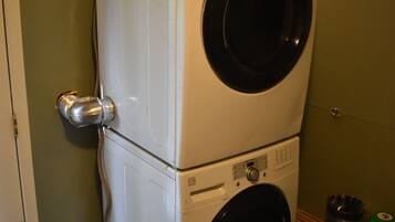 A stacked washer and dryer in a room with green walls, a wooden shelf, and a trash bin.