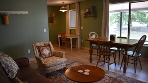 A living room with a brown sofa, patterned armchair, and a round coffee table with three white objects on it. There is a dining area with a wooden table and chairs, and a window with a view of trees. - Wildflower Suites North (Garfield)