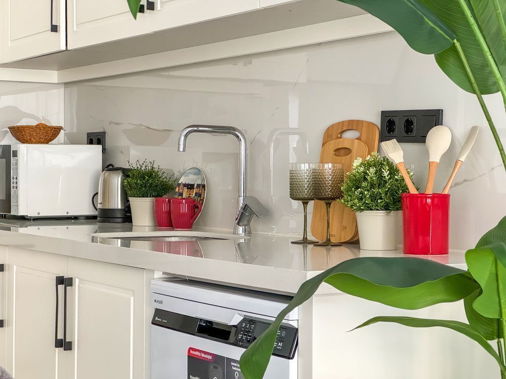 Uma cozinha moderna com armários brancos, uma pia de aço inoxidável e uma máquina de lavar louça. Há plantas em vasos e utensílios de cozinha no balcão.