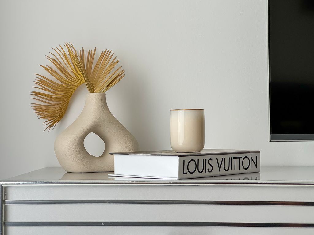 Eine weiße Vase mit außergewöhnlicher Form und eine beige Tasse auf einem weißen Buch mit der Aufschrift "LOUIS VUITTON" auf einer weißen Oberfläche.