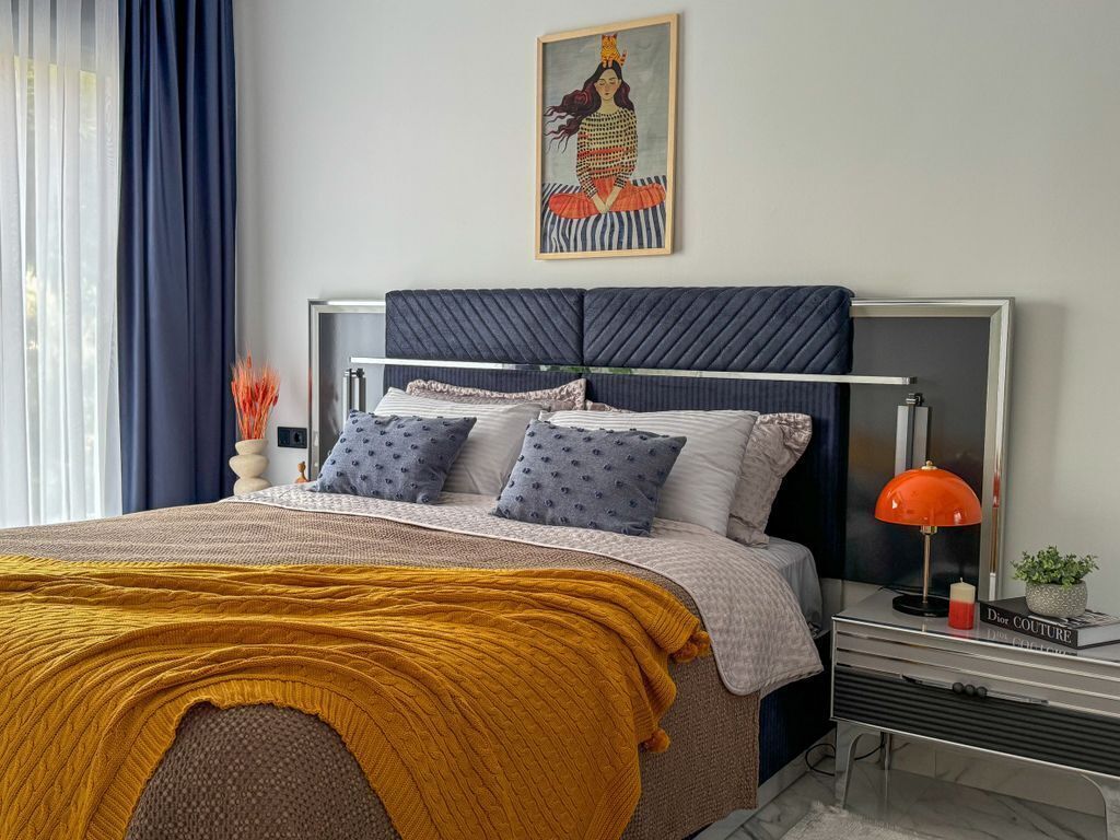 A modern bedroom with a bed, a bedside table with a lamp, a framed artwork on the wall, and a window with curtains.