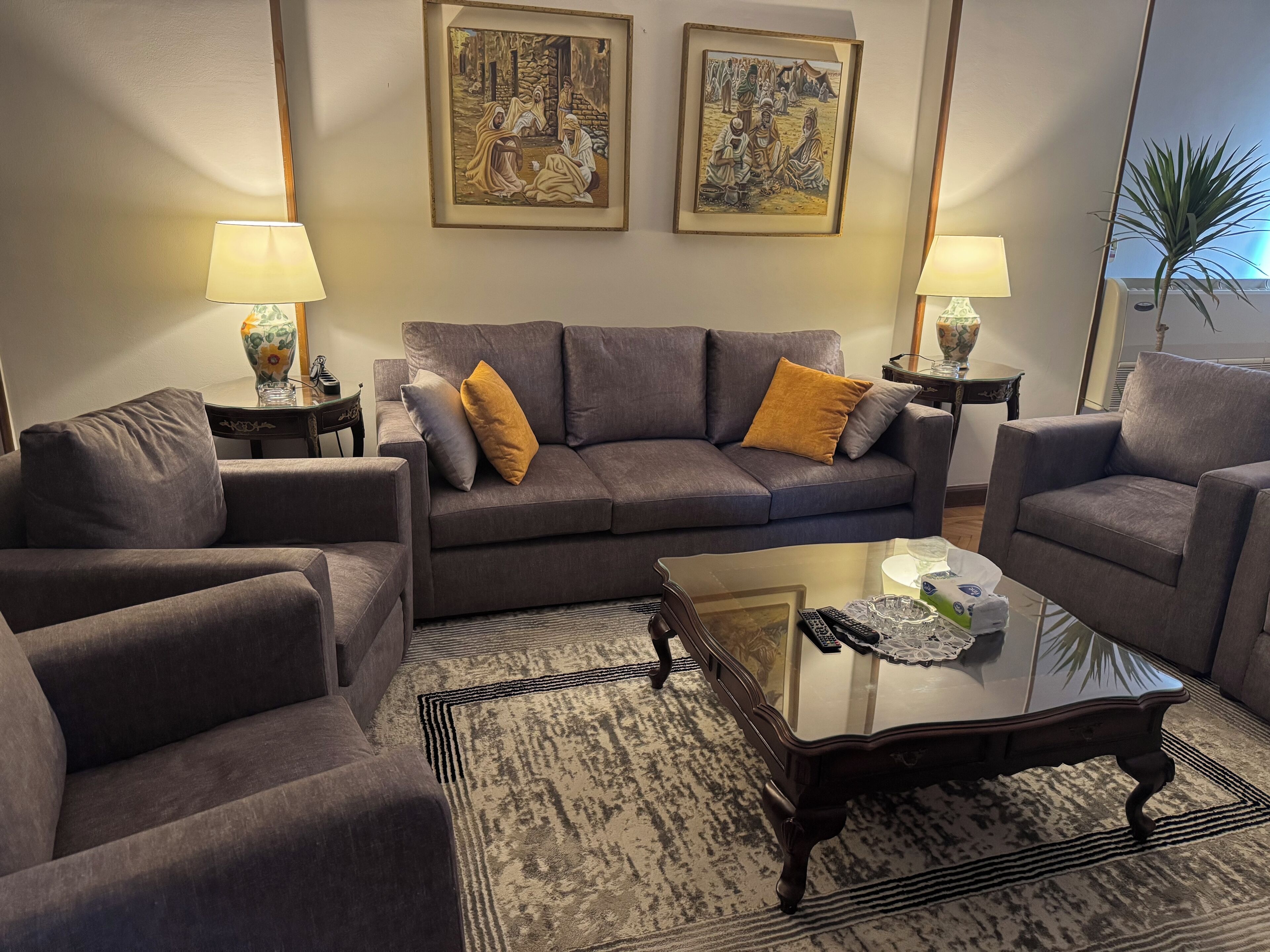 Ein modernes Wohnzimmer mit einem grauen Sofa, zwei Sesseln, einem Glastisch und einem gemusterten Teppich. Auf Beistelltischen stehen zwei Lampen und an der Wand hängen zwei gerahmte Kunstwerke.