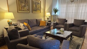 Ein modernes Wohnzimmer mit einer grauen Sofagarnitur, zwei Sesseln, einem Couchtisch und zwei Lampen. An der Wand befinden sich gerahmte Kunstwerke und darüber ein Kronleuchter.