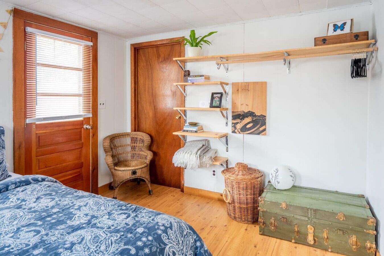 Ein Schlafzimmer mit einem Bett, einem Sessel, einer Holztür, einem Regal mit Dekoration und einem grünen Truhenkasten.